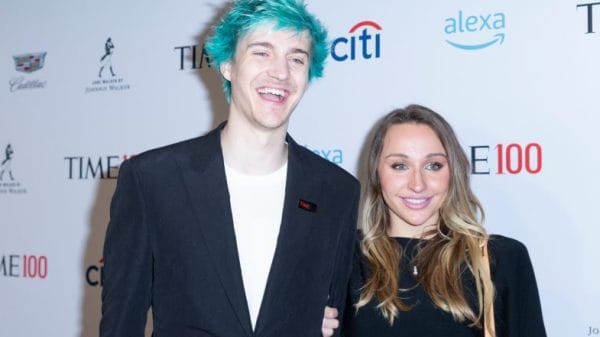 Ninja, a white man with blue hair in a black sit jacket and white tee, stands next to his wife, a shorter blonde woman in a black dress at an event