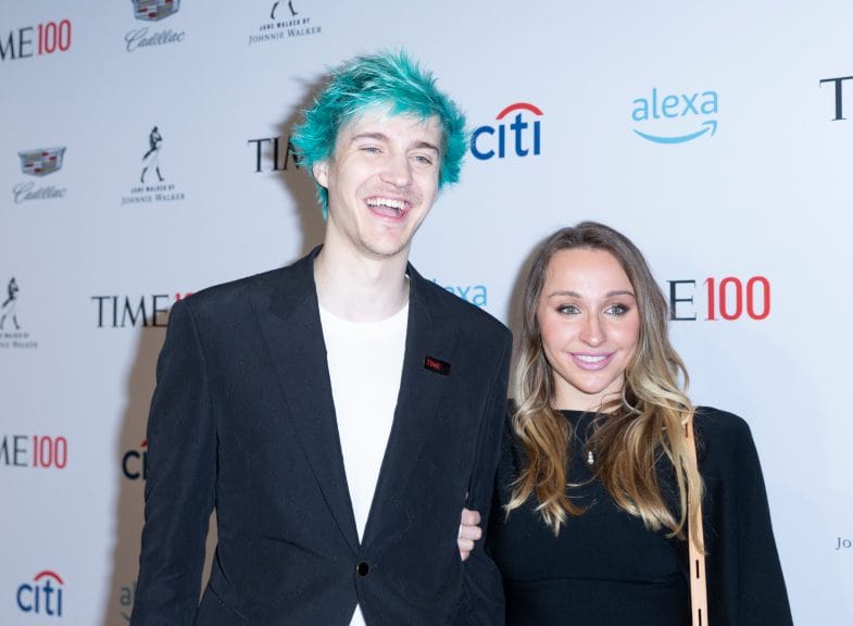 Ninja, a white man with blue hair in a black sit jacket and white tee, stands next to his wife, a shorter blonde woman in a black dress at an event