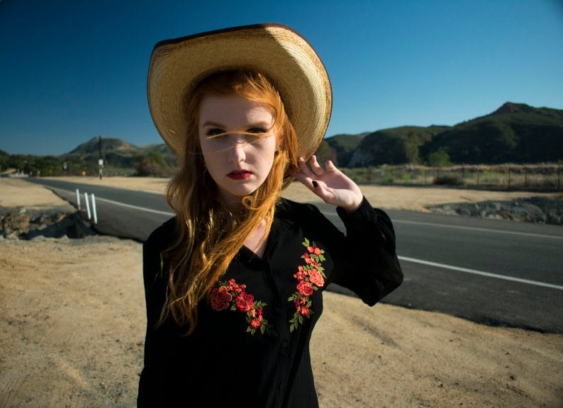 Redhead girl in black shirt and cowboy hat standing in front of a desert highway.