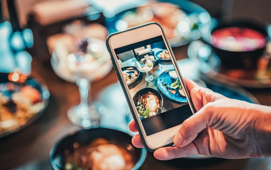 Focus on a phone, that is taking a photo of a table full of food.