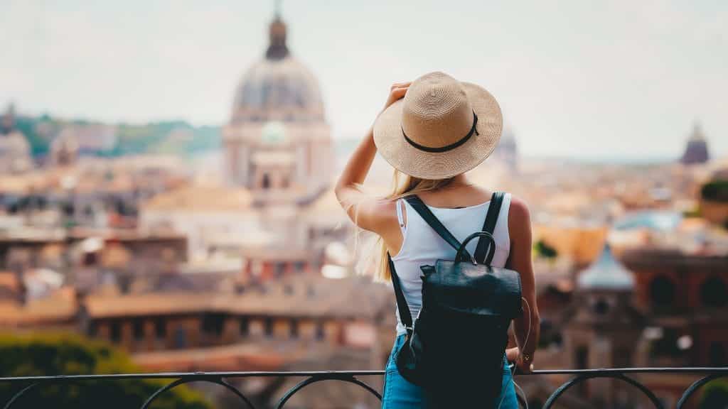 A young woman standing outside, looking around in front of the fence. Travelling is an excellent way to celebrate.