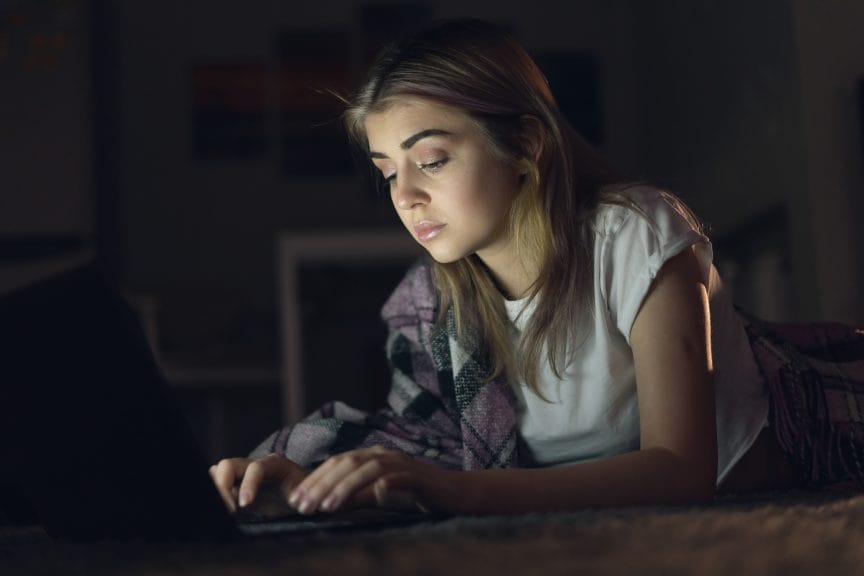 teenage girl watching a video or working on her laptop at night in the dark. The laptop screen lights up her face. 