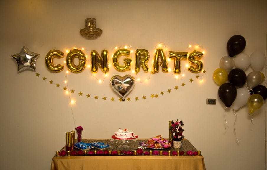 A white wall with balloons on it that says "Congrats" and a table with decorations and food on it. 
