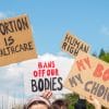 Four signs are up in the air reading: "abortion is healthcare" "bans off our bodies" "human right" "my body my choice"