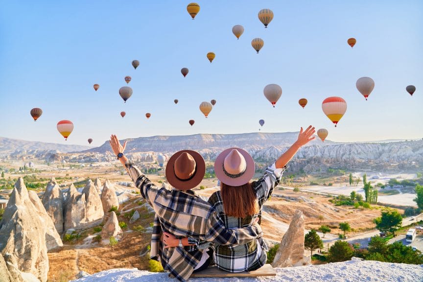 Two people sat on a cliff edge watching several hot air balloons in the sky.