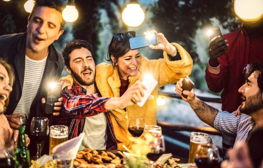 A group of young adults, are outside, sitting around a table with food and all have their phones out with their flashlights on.