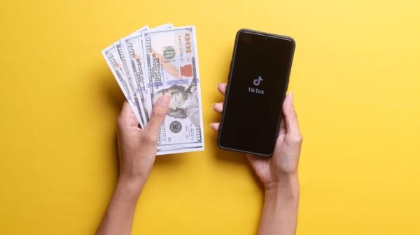 top down view of two hands against a yellow background. one hand has cash, the other has a phone with the tiktok logo.
