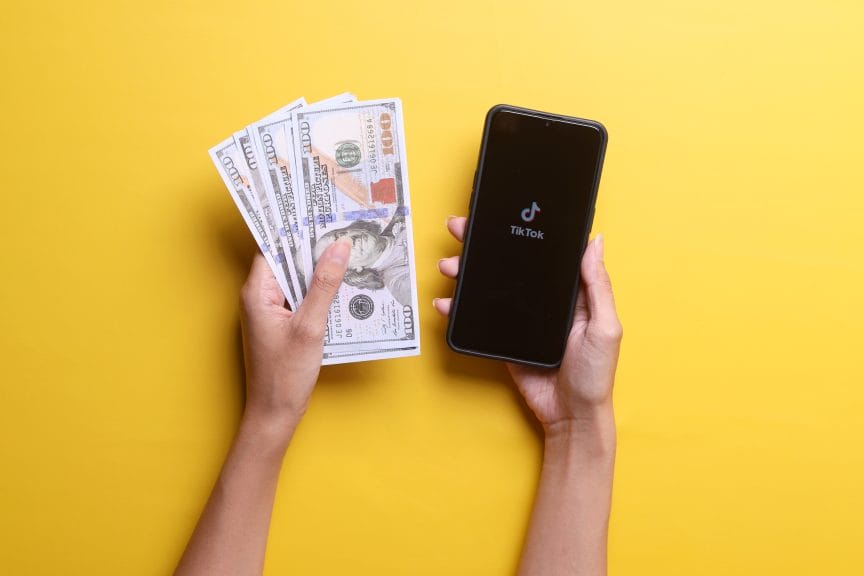 top down view of two hands against a yellow background. one hand has cash, the other has a phone with the tiktok logo.