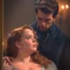 A red-haired woman and dark-haired man stand in front of a mirror making intense eye contact.