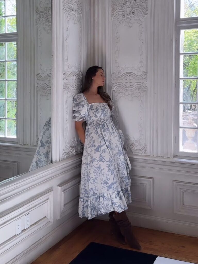 A woman wearing a floral white and blue dress, leaned against a corner in her house.