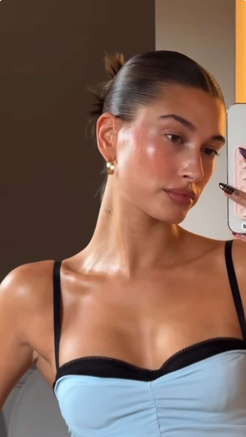 Hailey Bieber taking a mirror selfie with glossed skin, a slick-back bun, and small gold hoops.