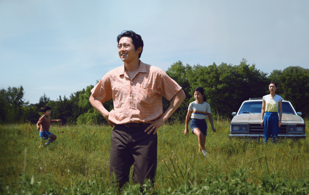 A family man is looking out into the fields as his family is behind him. The kids are playing in the fields and the wife by the car.