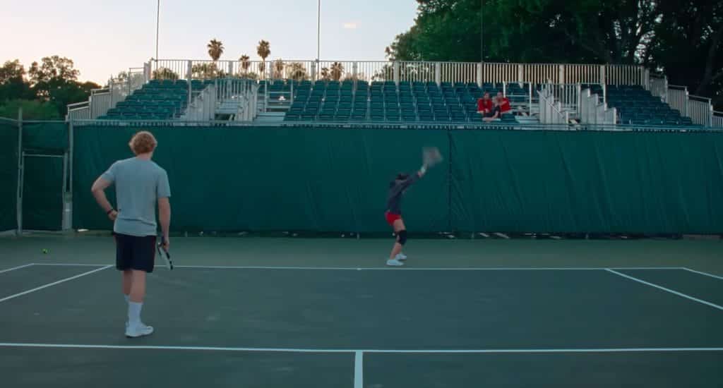 In the foreground, a man holding a tennis raquet, in the background, a woman smashing a racquet.