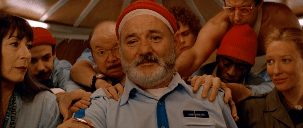 A group of people laying their hands on the shoulders of a man in the centre of the group. Some are wearing red beanies. They are in a submarine.
