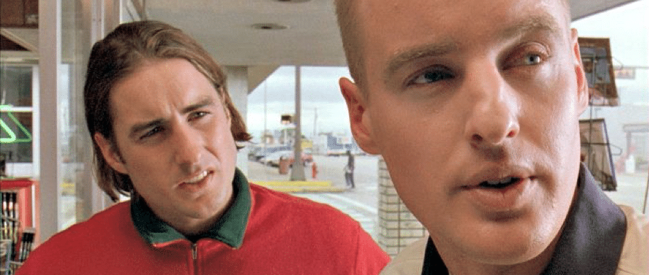 A man with long, brown hair in a red sweater in the background and a man in a white shirt in the foreground staring at something.