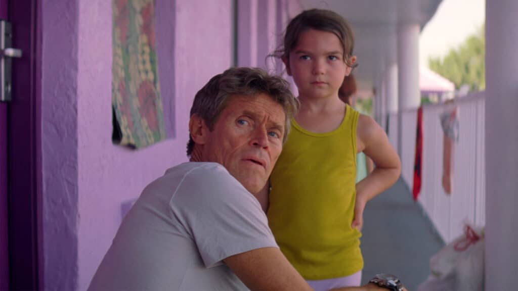 An older man is sitting crouched in front of a little girl, but both are looking behind the mans back at something. They both look serious.