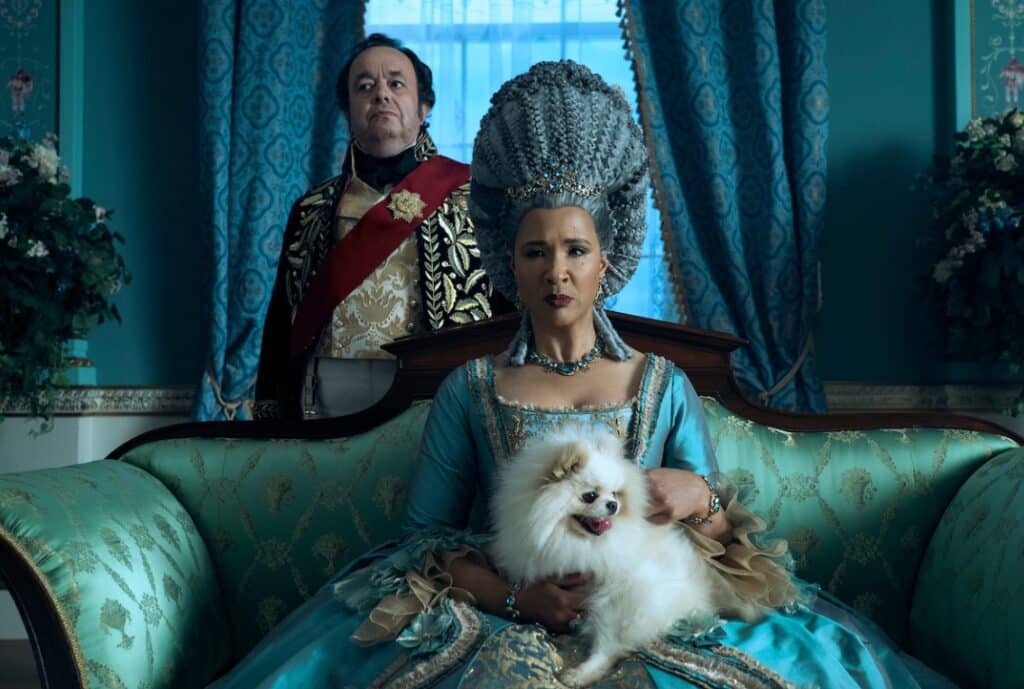 Queen Charlotte from Bridgerton sitting in an elegant dress and wig with her pet dog in her lap.