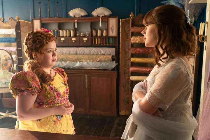 Eloise, a young white, dark haired woman wearing a light blue dress, stands face-to-face with Penelope, a young, white, red-haired woman wearing a yellow dress. They look at each other with serious expressions.