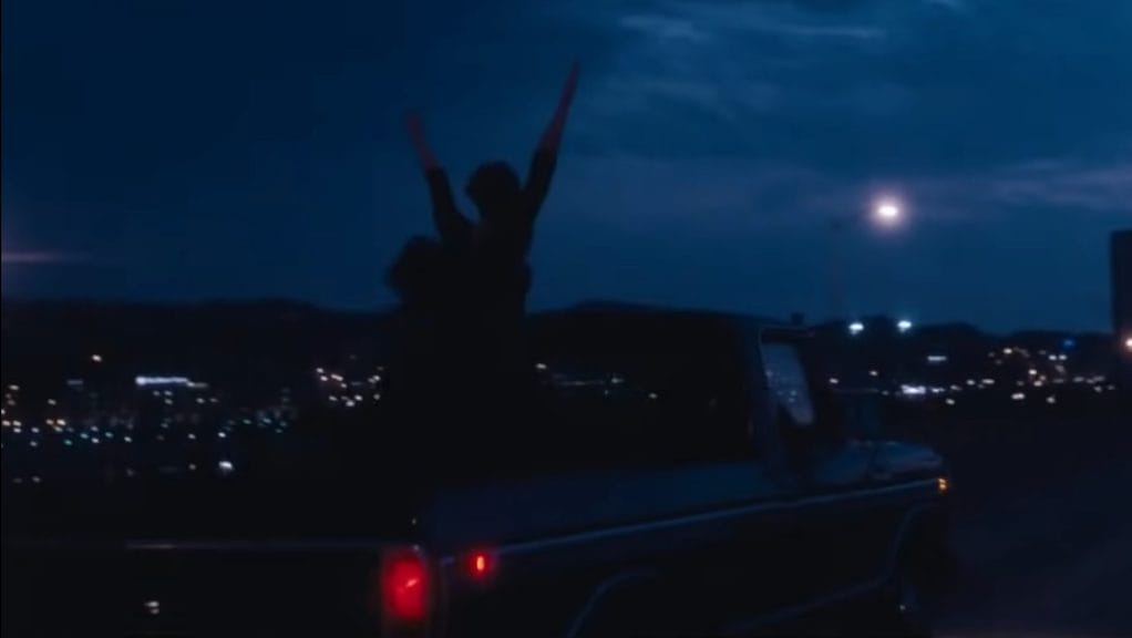 Sam, from The Perks of Being a Wallflower, stands against the backdrop of a dark night in the bed of a truck with her arms in the air.