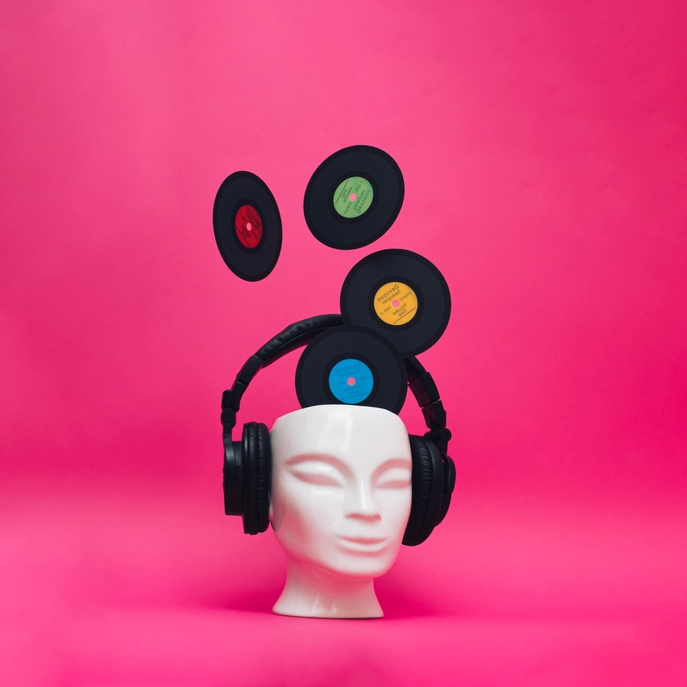 A mannequin head wearing headphones with vinyl records emerging from the top of the head. Pink background. Contemporary art with a retro music concept.
