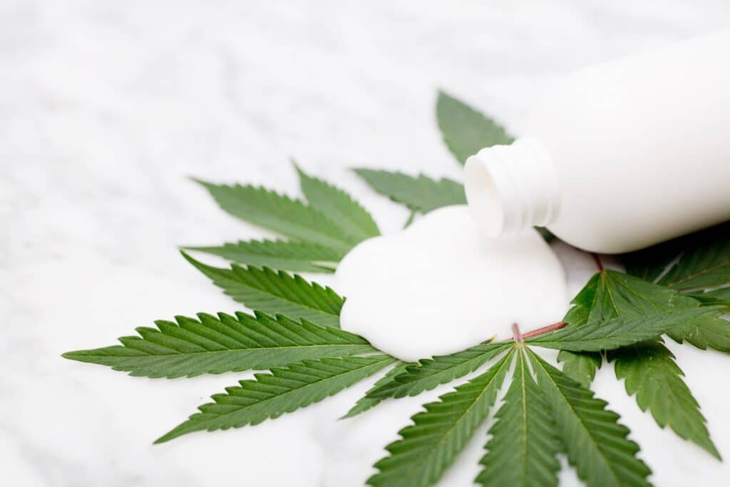 A white bottle lies on its side on a white surface. A white liquid is spilling out of the bottle and covering cannabis leaves.