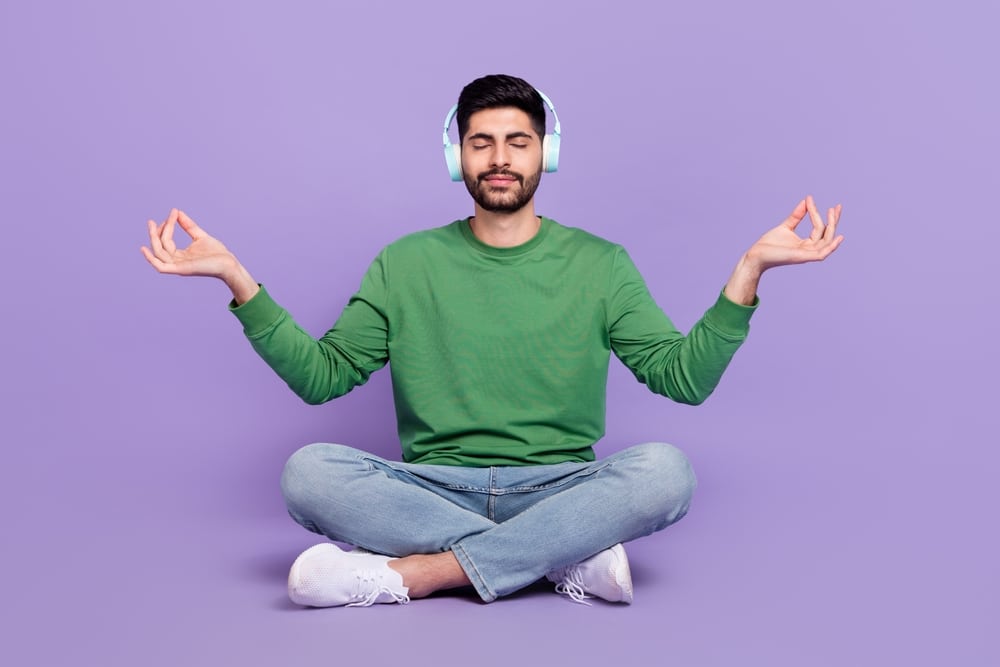 A man listening to music while meditating in the lotus pose. Appears to be in a good mood. Purple background.