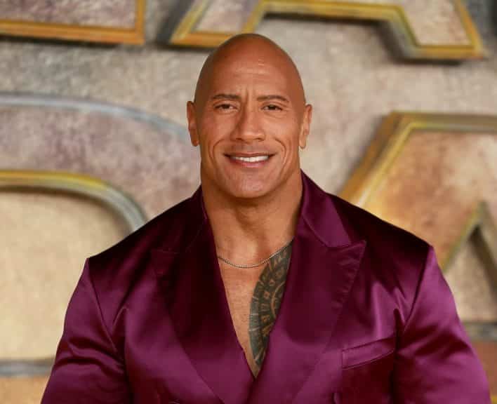 A bald Samoan man in a purple silk suit stands smiling in front of the words 'Black Adam'
