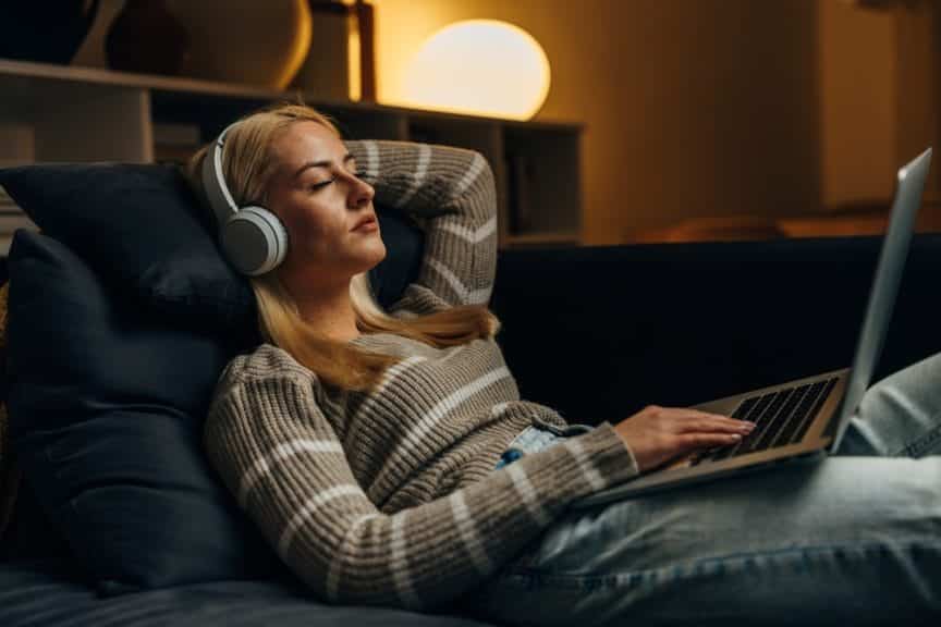 Blonde woman listening to music on social media while sitting on a couch with headphones and laptop