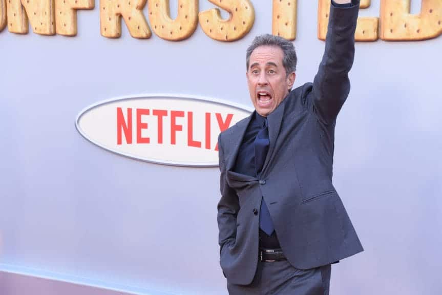 A man with grey hair in a grey suit with one arm in the hair stands with his mouth open with the netflix logo in the background