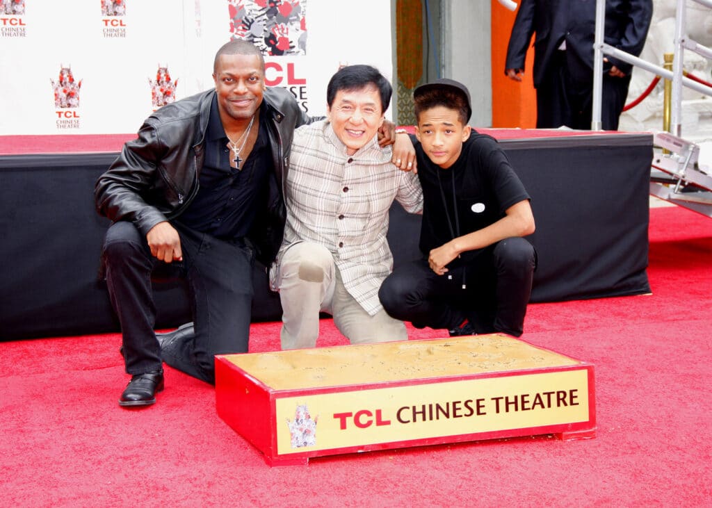 A black man in a leather jacket and black trousers crouches down next to an asian man in a biege suit and a black teenager in a black-tshirt and trousers behind a white backdrop on a red carpet with a yellow platform on the ground that reads 'TCL Chinese Theatre'