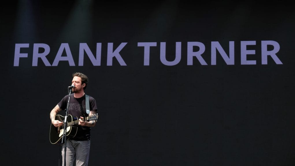 Frank Turner is singing onstage at Victorious Festial, against a black backdrop with his name in blue