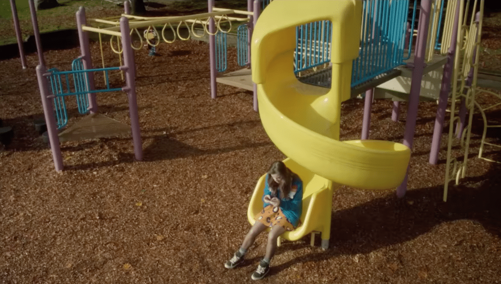 Nadine, the main character of the movie The Edge of Seventeen sits on her phone at the bottom of a swirly slide. She is at a playground.