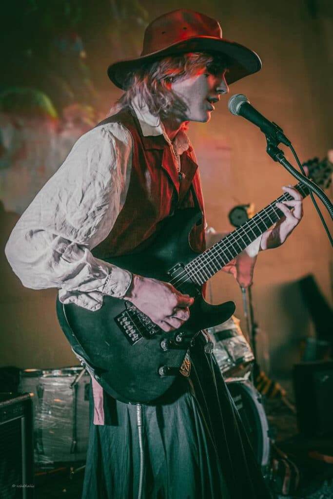A musician on stage, singing into a mic while playing a black guitar.
