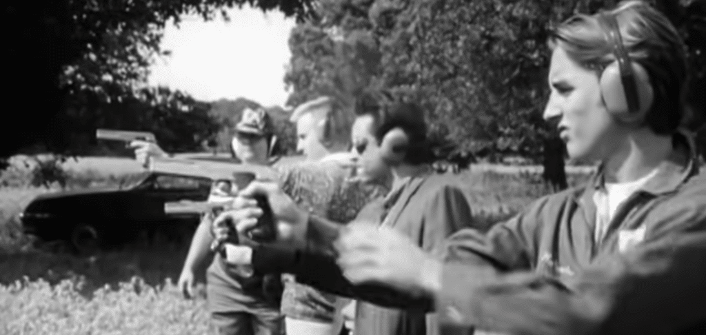 Four men in a row in the middle of a field, pointing guns. The image is in black and white.