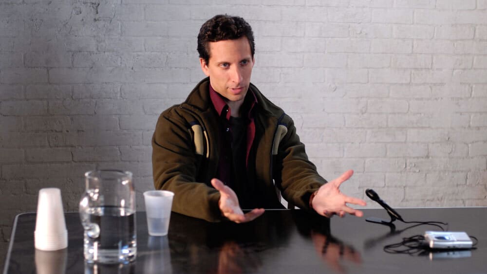 A man at a table with a microphone on his left and a water jug on his right. He is gesturing with his hands, as if explaining something.