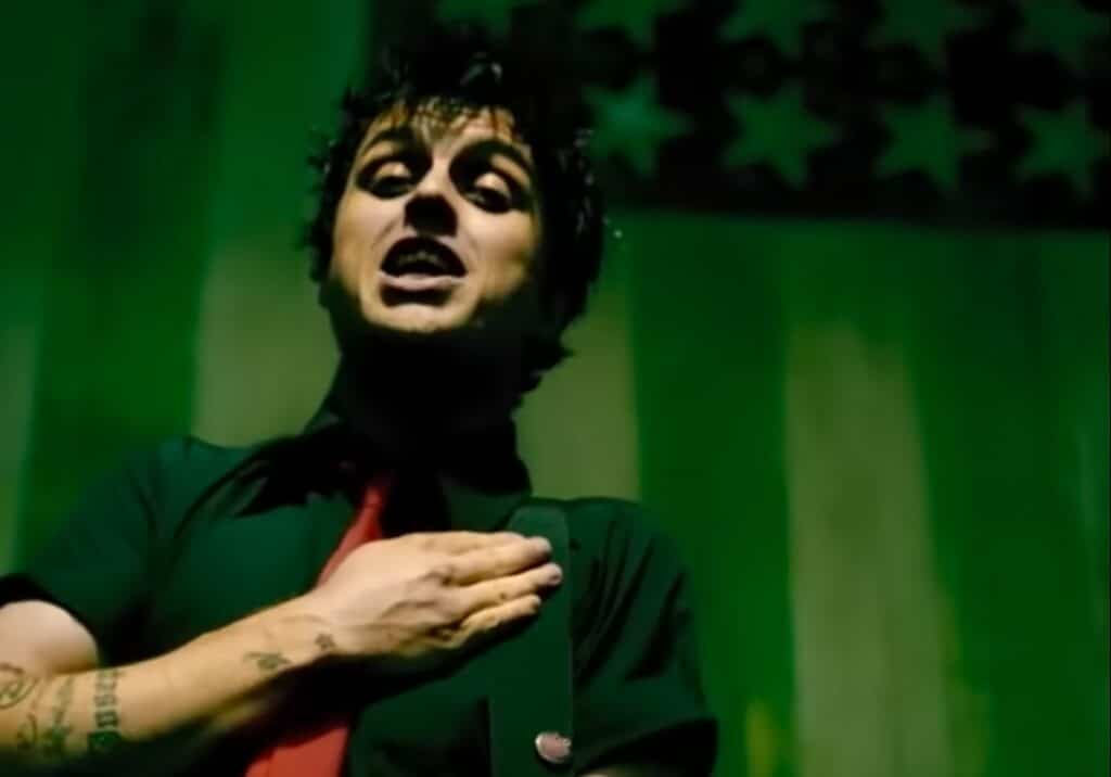 Billie Joe Armstrong with his hand over his heart. Behind him, there is a green version of the United States flag.