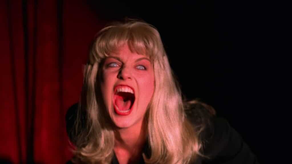 A blonde woman screaming. Behind her is a red curtain. Her eyes are glassy.