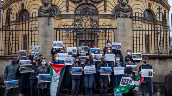 students with faces covered and wearing blac stand holding pictures of and information about destroyed education buildings