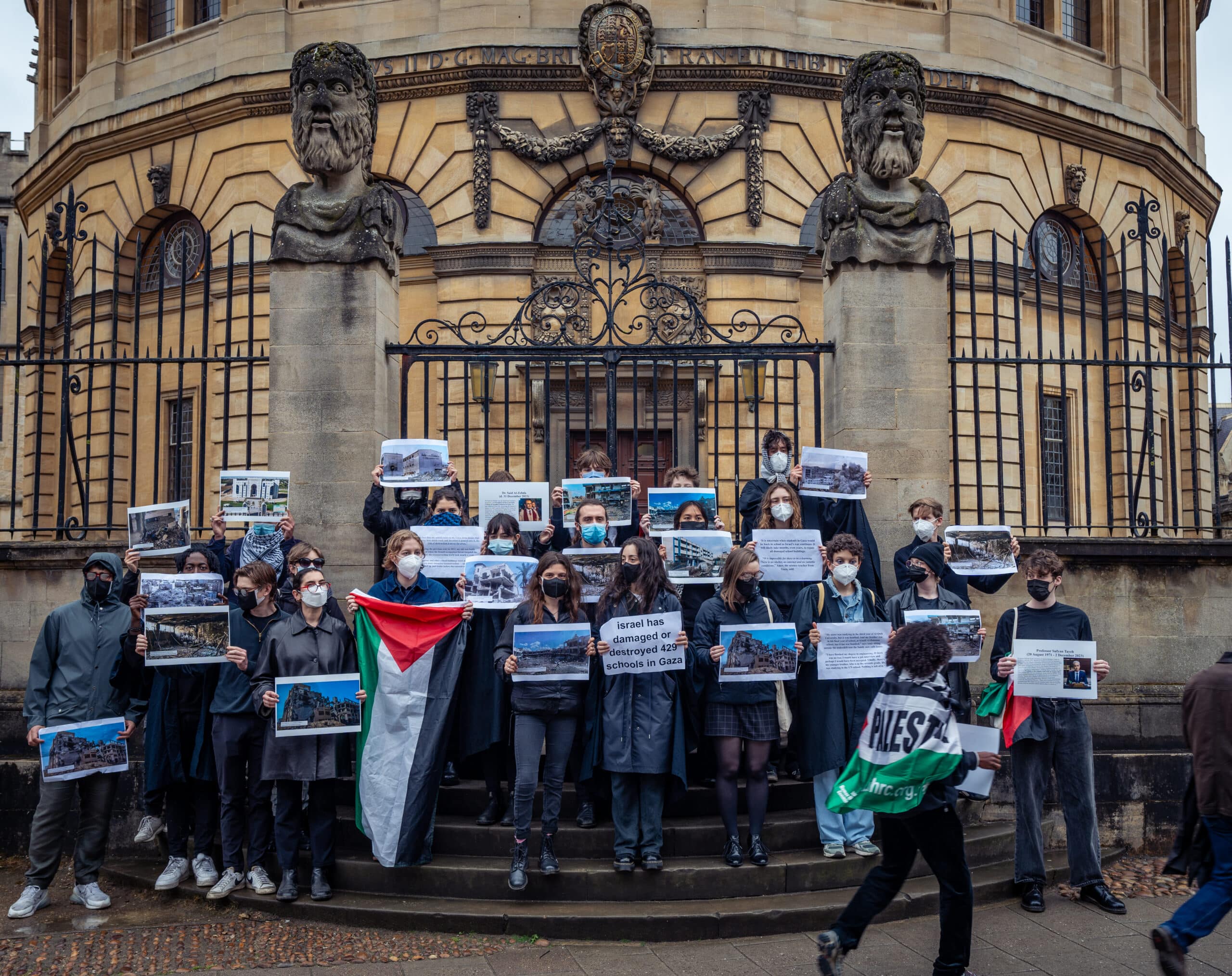 students with faces covered and wearing blac stand holding pictures of and information about destroyed education buildings
