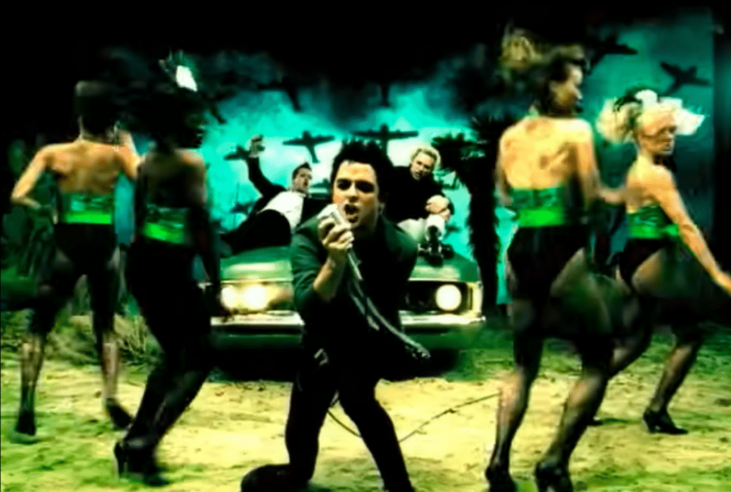 Billie Joe Armstrong is singing into a microphone with backup dancers. Behind them are Tré Cool and Mike Dirnt lying on a car.