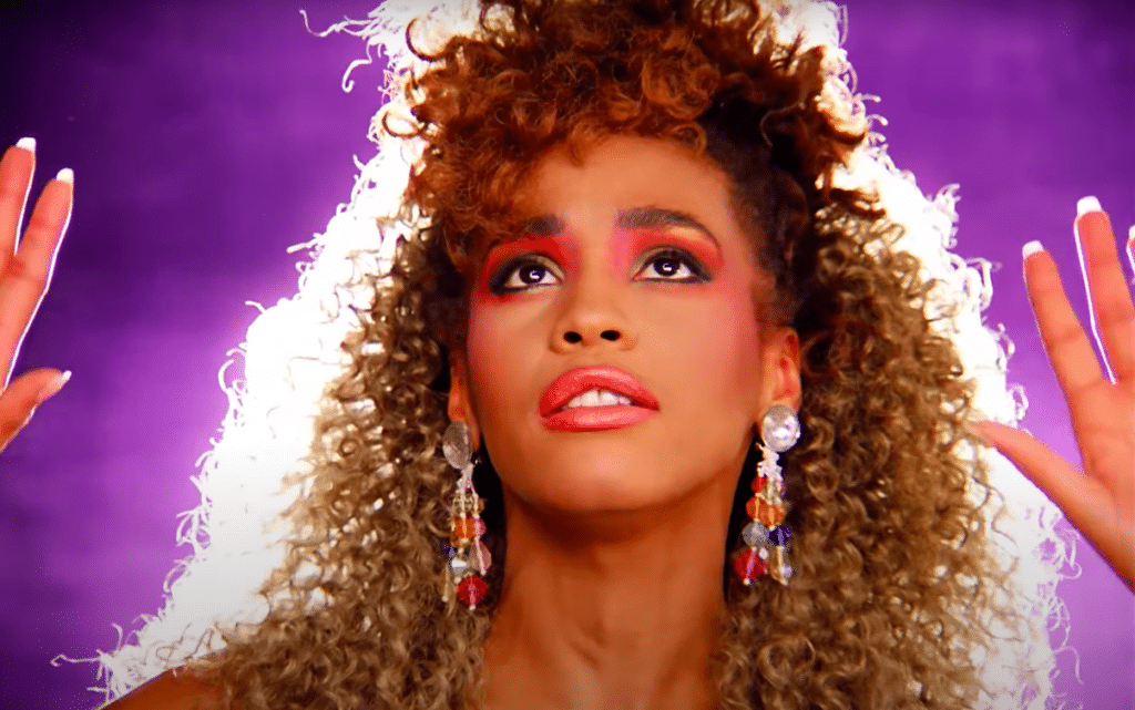 Whitney Houston is in front of a purple backdrop looking up with her hands next to her face.