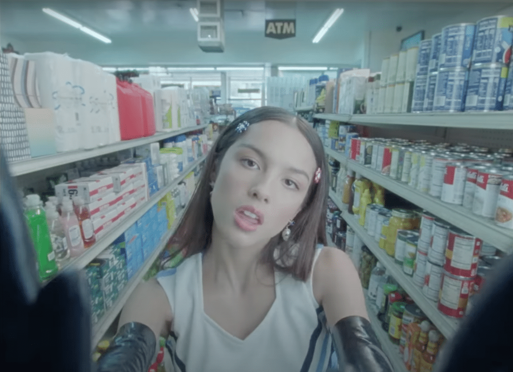 Olivia Rodrigo in the middle on a grocery aisle with both hands on pressed against the bottom side lens of camera