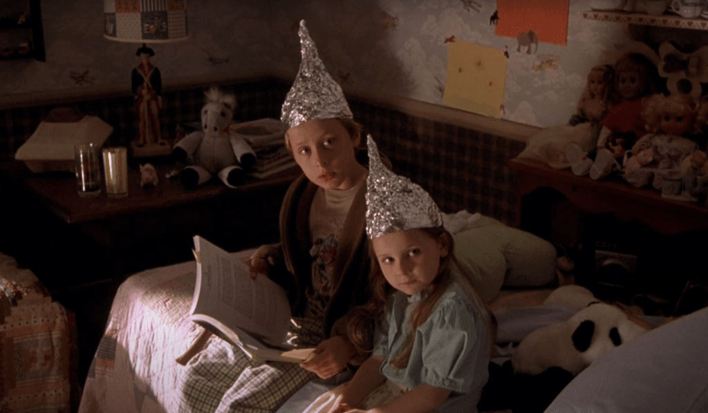 A boy and a girl are sitting on the bed wearing aluminum hats.