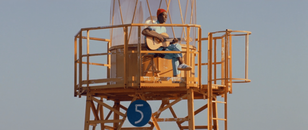 A man playing guitar atop a tower made of yellow scaffolding. He is in blue coveralls and a red hat.