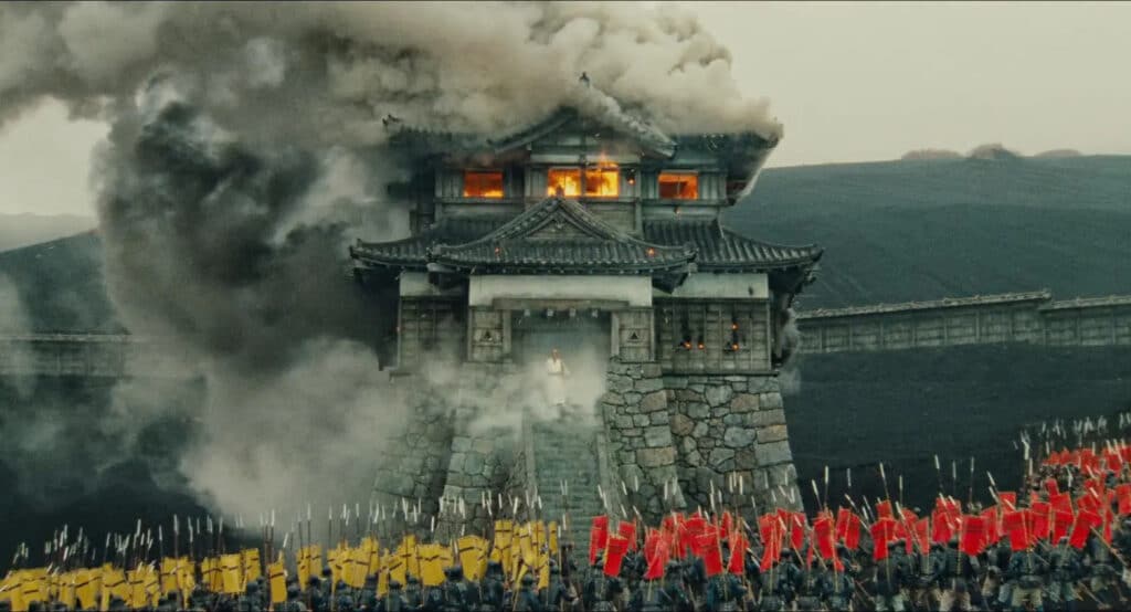 A battle halts before a burning castle, watching Hidetora (Tatsuya Nakadai) descends the steps.