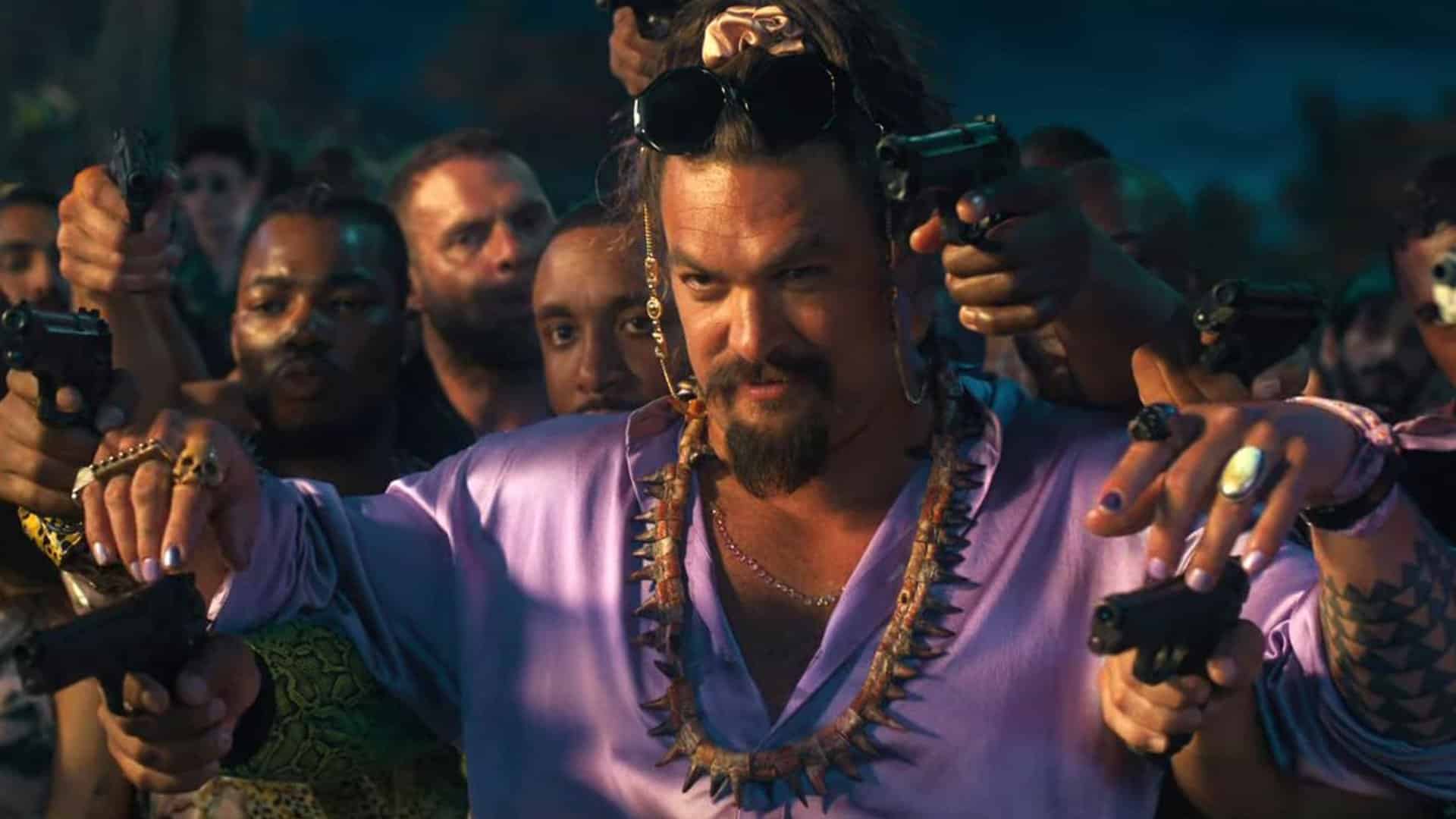 A tough-looking mand wearing a purple button down shirt gloats to somebody as many others point guns over his shoulders towards the person he talks to.