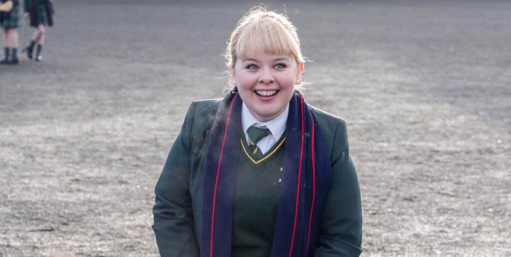 A young, blond white girl with blunt bangs, wearing a green school uniform smiles broadly.