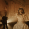 Chappell Roan wears a wedding dress in an industrial set. Light shines behind her as she holds a microphone. Behind her are two guitarists.