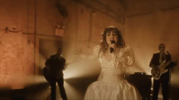 Chappell Roan wears a wedding dress in an industrial set. Light shines behind her as she holds a microphone. Behind her are two guitarists.