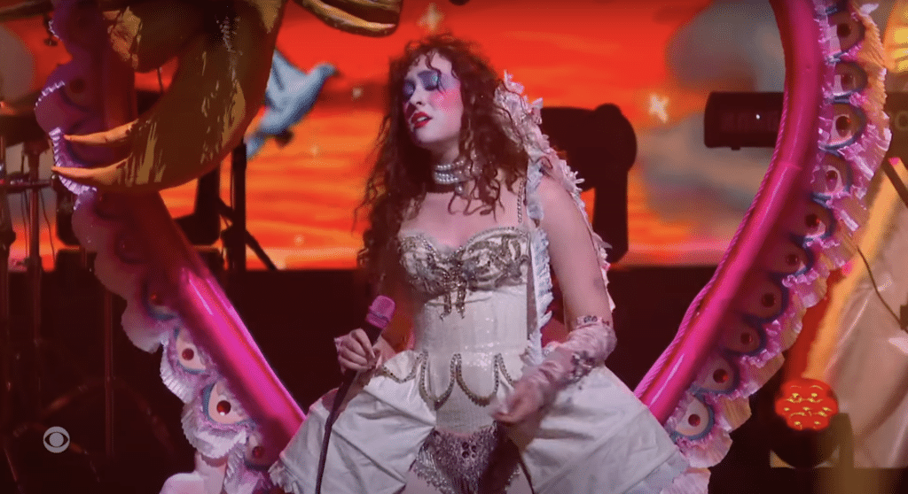 Chappell Roan performs in front of a heart-shaped prop. She is wearing a corset top with a Rococo-inspired petticoat.